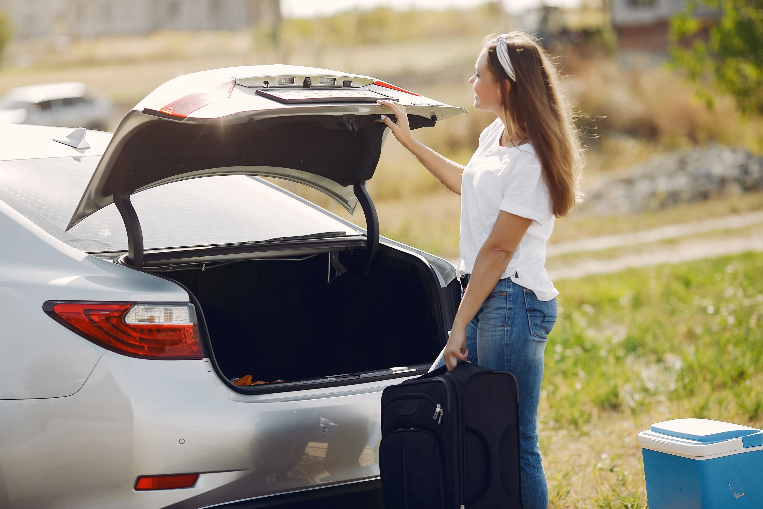 A woman holding a black suitcase opens the boot of a silver saloon car on a sunny day, with a blue cool box on the grass beside her