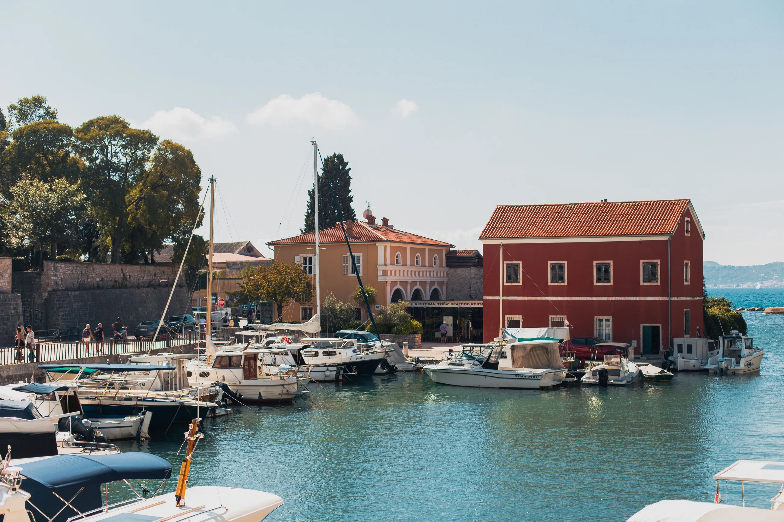 Motor boats and sailing boats moored in a small harbour in Zadar on a sunny day, with a red house at the water’s edge and a road with parked cars beside an old stone wall