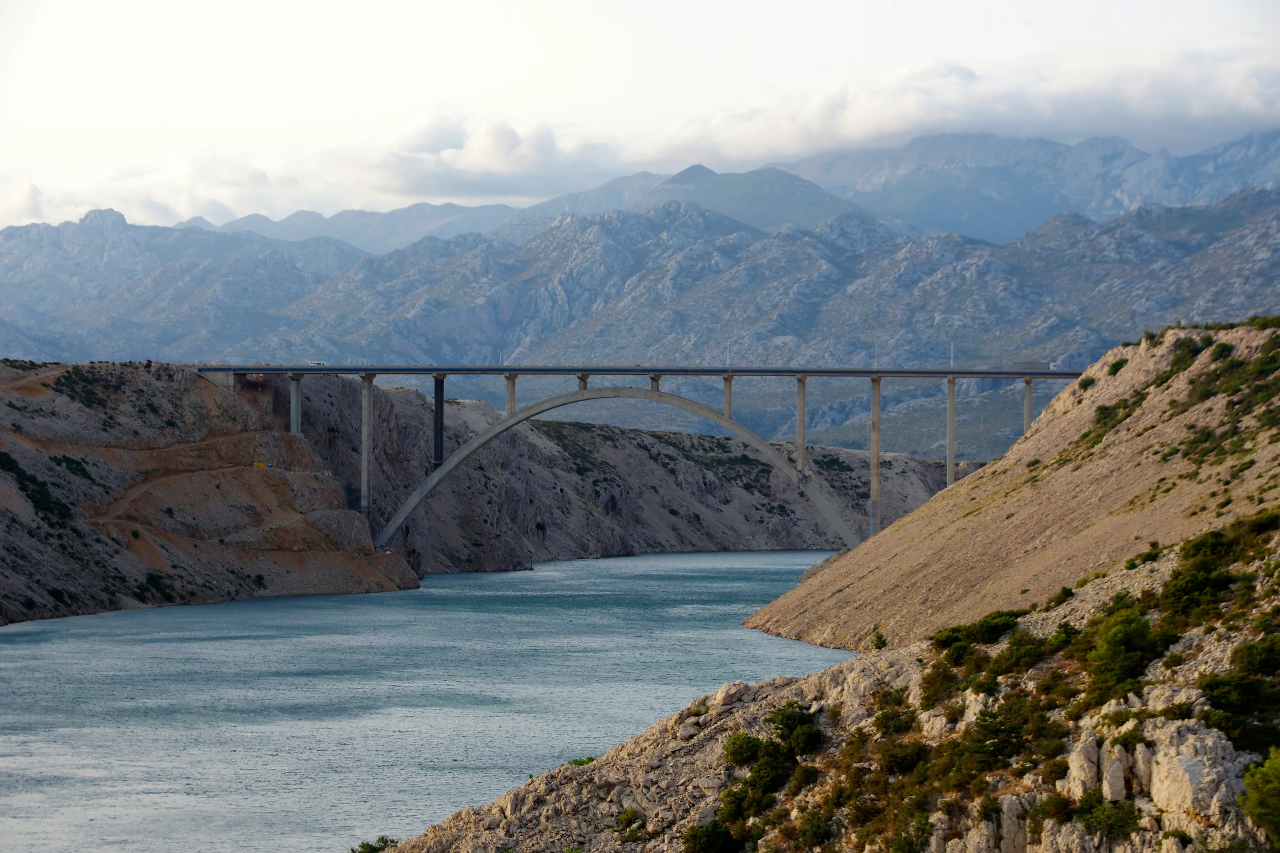 The Maslenica bridge: a concrete arch road bridge over a narrow channel of water between bare rocky slopes, with a mountain range capped in cloud behind it